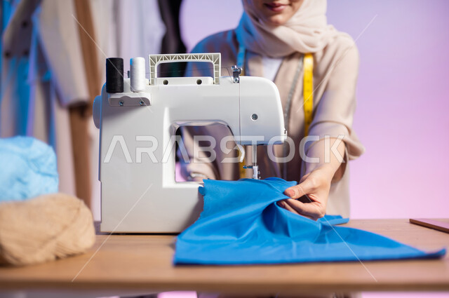 استخدام ماكينة الخياطة لتفصيل الملابس النسائية , مهن و حرف يدوية في السعودية , بورتريه مقرب لمصممة أزياء عربية خليجية سعودية ترتدي الزي الكاجوال و الحجاب , ممارسة مهنة الخياطة , خلفية ملونة