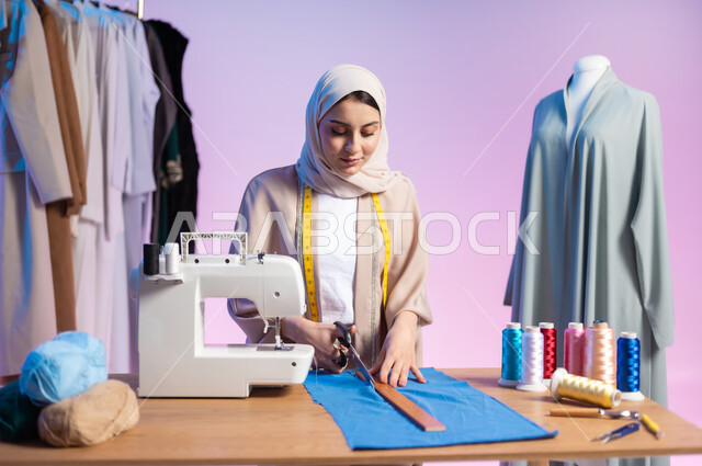 استخدام المسطرة و المقص في تفصيل الملابس , ممارسة مهنة الخياطة , بورتريه مقرب لمصممة أزياء عربية خليجية سعودية ترتدي الزي الكاجوال و الحجاب تقوم بقص قماشة لتفصيل ملابس نسائية , خلفية ملونة