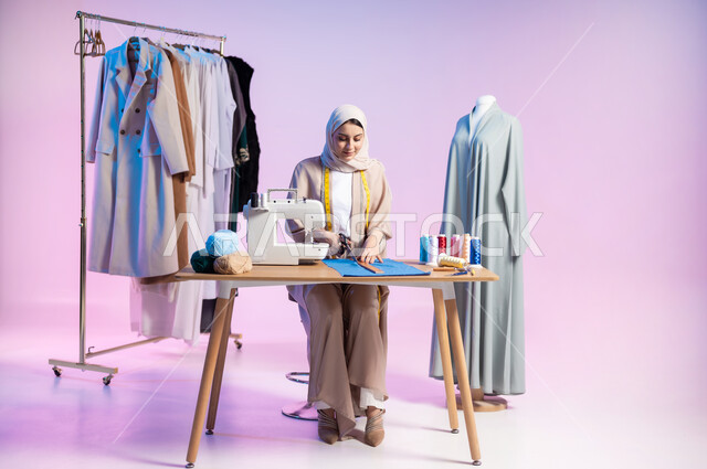 استخدام الأدوات المناسبة للحياكة , ممارسة مهنة الخياطة , بورتريه لمصممة أزياء عربية خليجية سعودية ترتدي الزي الكاجوال و الحجاب تقوم بقص قماشة لتفصيل ملابس نسائية , صورة للجسم بالطول الكامل , خلفية ملونة