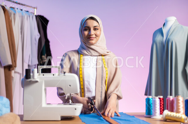 حرف و مهن يدوية , ممارسة مهنة الخياطة , بورتريه مقرب لمصممة أزياء عربية خليجية سعودية ترتدي الزي الكاجوال و الحجاب تقوم بقص قماشة لتفصيل ملابس نسائية , النظر إلى الكاميرا بإيماءات الفرح و السرور , خلفية ملونة