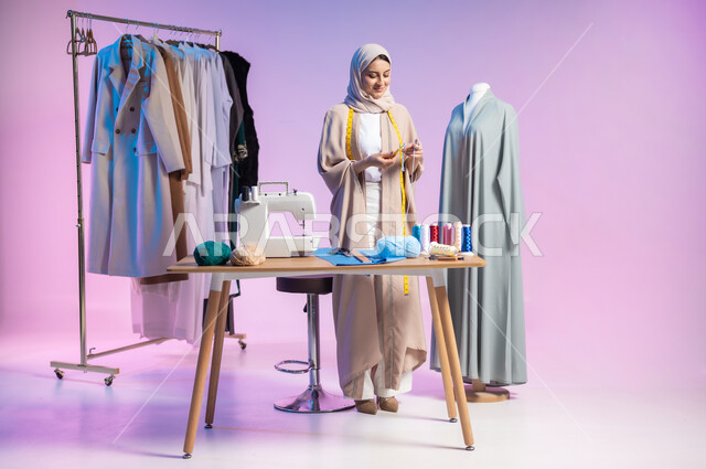 إتقان صناعة الملابس السعودية، ممارسة مهنة الحياكة، استعمال أدوات ومعدات الخياطة، بورتريه لمصممة أزياء عربية خليجية سعودية ترتدي الزي الكاجوال والحجاب تمسك بيدها السنارة والصوف, اختيار أجود أنواع الأقمشة لصنع الثياب، الجسم بالطول الكامل، خلفية ملونة