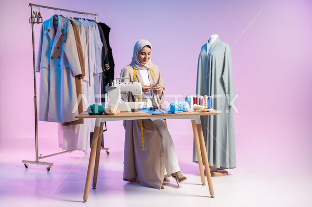 ممارسة مهنة الحياكة، استعمال أدوات ومعدات الخياطة، بورتريه لمصممة أزياء عربية خليجية سعودية ترتدي الزي الكاجوال والحجاب تمسك بيدها السنارة والصوف, إتقان صناعة الملابس السعودية، اختيار أجود أنواع الأقمشة لصنع الثياب، الجسم بالطول الكامل، خلفية ملونة