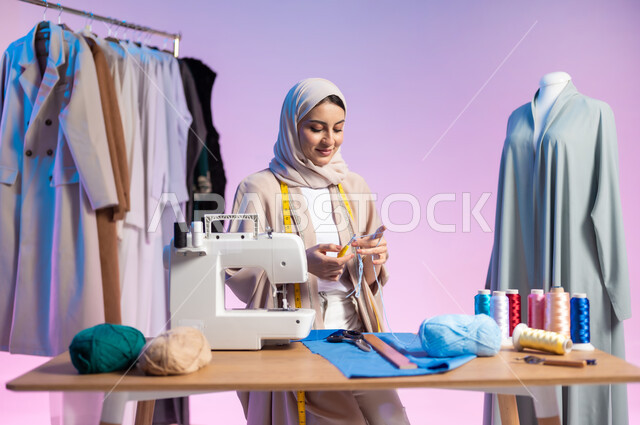 حرف ومهن يدوية، استعمال أدوات ومعدات الخياطة، بورتريه مقرب لمصممة أزياء عربية خليجية سعودية ترتدي الزي الكاجوال والحجاب تمسك بيدها السنارة والصوف, إتقان صناعة الملابس السعودية، اختيار أجود أنواع الأقمشة لصنع الثياب، خلفية ملونة