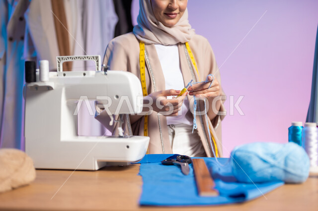 استعمال أدوات ومعدات الخياطة، حرف ومهن يدوية، بورتريه مقرب لمصممة أزياء عربية خليجية سعودية ترتدي الزي الكاجوال والحجاب تمسك بيدها السنارة والصوف, إتقان صناعة الملابس السعودية، اختيار أجود أنواع الأقمشة لصنع الثياب، خلفية ملونة