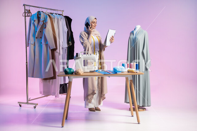 إجراء مكالمة هاتفية، استخدام التكنولوجيا المتطورة، بورتريه لمصممة أزياء عربية خليجية سعودية ترتدي الزي الكاجوال والحجاب تستخدم الهاتف المحمول, إتقان صناعة الملابس السعودية، اختيار أجود أنواع الأقمشة لصنع الثياب، الجسم بالطول الكامل، خلفية ملونة