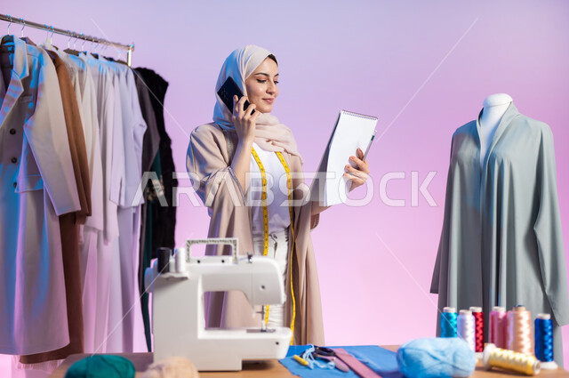 Using advanced technology, making a phone call, a close-up portrait of a Saudi Arabian Gulf fashion designer wearing a casual outfit and hijab, using a mobile phone, mastering the Saudi clothing industry, choosing the finest types of fabrics to make clothes, using sewing tools, a colorful background