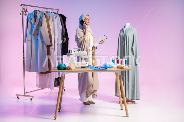 استخدام التقنيات الحديثة، إتقان صناعة الملابس السعودية، اختيار أجود أنواع الأقمشة لصنع الثياب، بورتريه لمصممة أزياء عربية خليجية سعودية ترتدي الزي الكاجوال والحجاب تستخدم الهاتف, إجراء مكالمة هاتفية، استعمال أدوات الخياطة، الجسم بالطول الكامل، خلفية ملونة