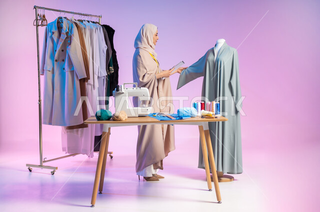 حرف ومهن يدوية، إتقان صناعة الملابس السعودية، اختيار أجود أنواع الأقمشة لصنع الثياب، بورتريه لمصممة أزياء عربية خليجية سعودية ترتدي الزي الكاجوال و الحجاب تمسك بيدها الدفتر والقلم, استعمال أدوات ومعدات الخياطة، الجسم بالطول الكامل، خلفية ملونة