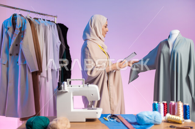 إتقان صناعة الملابس السعودية، اختيار أجود أنواع الأقمشة لصنع الثياب، بورتريه مقرب لمصممة أزياء عربية خليجية سعودية ترتدي الزي الكاجوال و الحجاب تمسك بيدها الدفتر والقلم, استعمال أدوات ومعدات الخياطة، حرف ومهن يدوية، خلفية ملونة