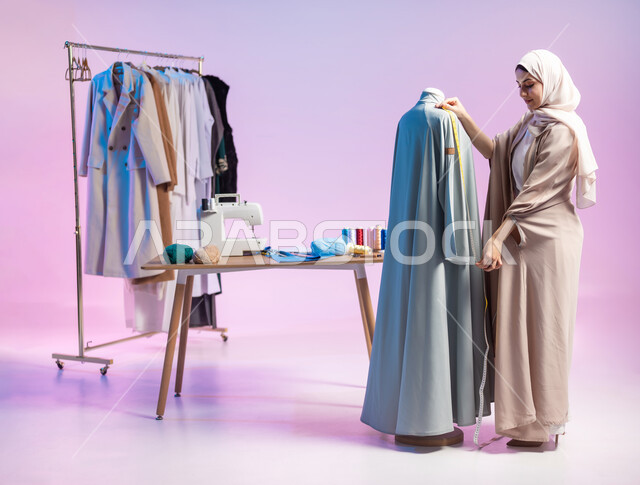 اختيار أجود أنواع الأقمشة لصنع الثياب، أخذ القياسات باستخدام الشريط المتري، بورتريه لمصممة أزياء عربية خليجية سعودية ترتدي الزي الكاجوال و الحجاب, إتقان صناعة الملابس السعودية، استعمال أدوات ومعدات الخياطة، الجسم بالطول الكامل، خلفية ملونة