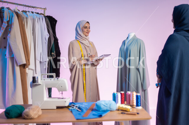 حرف ومهن يدوية، تدوين القياسات والملاحظات، إتقان صناعة الملابس السعودية، اختيار أجود أنواع  الأقمشة لصنع الثياب، بورتريه مقرب لمصممة أزياء عربية خليجية سعودية ترتدي الزي الكاجوال و الحجاب تحمل بيدها الدفتر والقلم, استعمال أدوات ومعدات الخياطة، خلفية ملونة