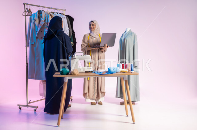 Mastering the Saudi clothing industry, using advanced technologies, choosing the finest types of clothing to make clothes, a portrait of a Saudi Arabian Gulf fashion designer wearing casual clothes and a hijab, holding a laptop in her hand, using sewing tools and equipment, full-length body, colored background