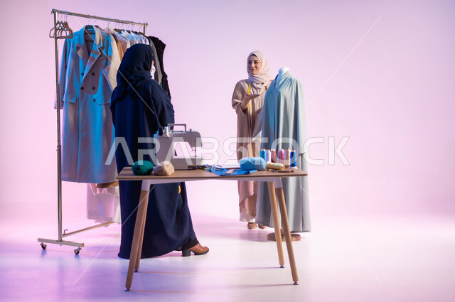 اختيار أجود أنواع  الأقمشة لصنع الثياب، ممارسة مهنة الحياكة, إتقان صناعة الملابس السعودية، بورتريه لمصممة أزياء عربية خليجية سعودية ترتدي الزي الكاجوال و الحجاب تشير بيدها الى المليكان, استعمال أدوات ومعدات الخياطة، الجسم بالطول الكامل، خلفية ملونة