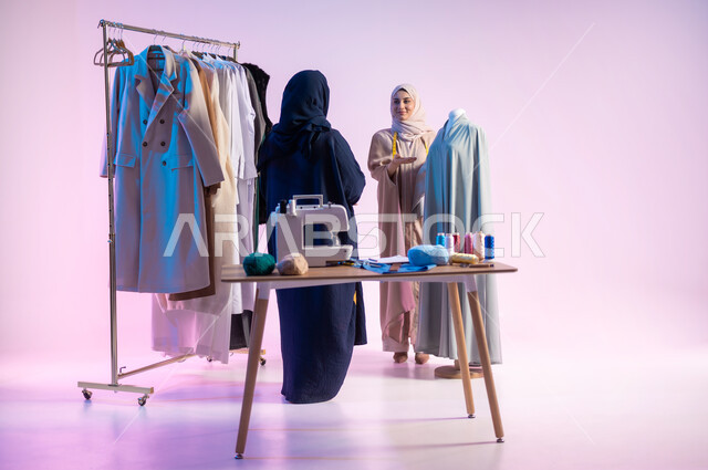 ممارسة مهنة الحياكة, إتقان صناعة الملابس السعودية، بورتريه لمصممة أزياء عربية خليجية سعودية ترتدي الزي الكاجوال و الحجاب تشير بيدها الى المليكان, استعمال أدوات ومعدات الخياطة، اختيار أجود أنواع  الأقمشة لصنع الثياب، الجسم بالطول الكامل، خلفية ملونة