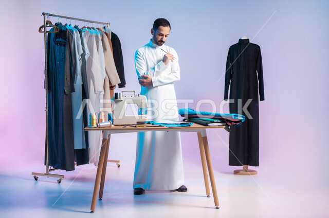 Using sewing tools and equipment, practicing the knitting profession, manual crafts and professions, mastering the manufacture of Saudi clothes, choosing the finest types of fabrics for making dresses, a portrait of a Saudi Arabian Gulf tailor wearing a traditional dress, holding a pen in his hand, full-length body, colored background.