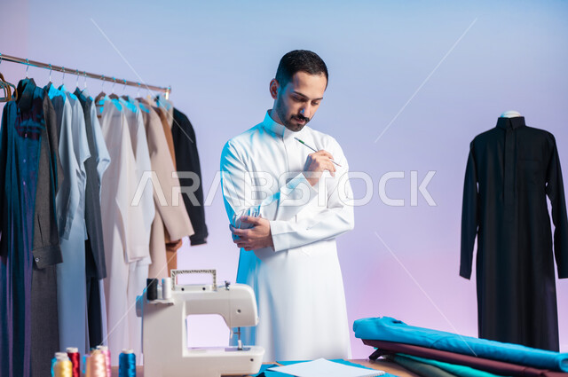 Practicing the profession of knitting, manual crafts and professions, mastering the manufacture of Saudi clothes, using sewing tools and equipment, choosing the finest types of fabrics for making dresses, a close-up portrait of a Saudi Arabian Gulf tailor wearing the traditional dress holding a pen in his hand, a colored background.