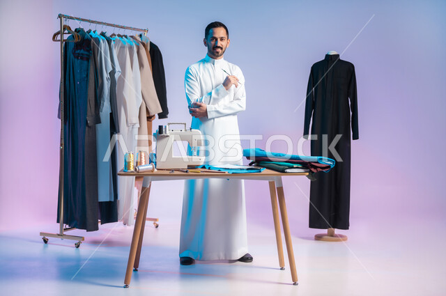 Looking at the camera with happy gestures, mastering making Saudi clothes, practicing the sewing profession, using sewing tools and equipment, choosing the finest types of fabrics for making dresses, portrait of a Saudi Arabian Gulf tailor wearing a traditional dress holding a pen in his hand, full length body, colored background
