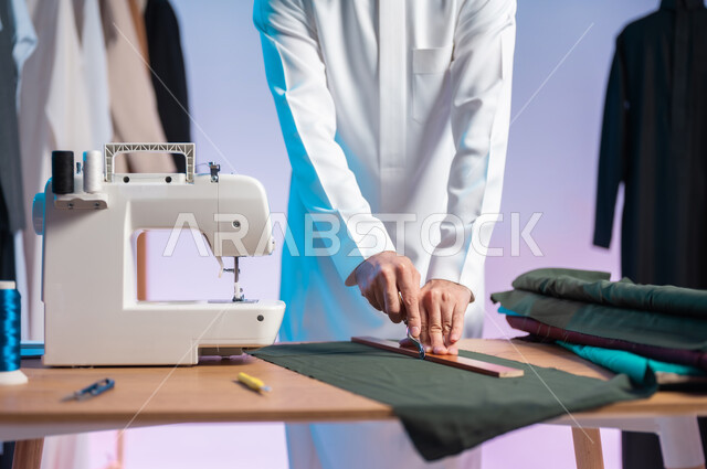 استعمال أدوات ومعدات الخياطة، اختيار أجود أنواع الأقمشة لصناعة الأثواب، إتقان صنع الملابس السعودية، حرف ومهن يدوية، بورتريه مقرب لخياط عربي خليجي سعودي يرتدي الثوب التقليدي يمسك المدرج والمسطرة، خلفية ملونة