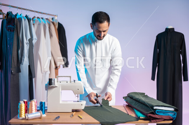 اختيار أجود أنواع الأقمشة لصناعة الأثواب، إتقان صنع الملابس السعودية، استعمال أدوات ومعدات الخياطة، حرف ومهن يدوية، بورتريه مقرب لخياط عربي خليجي سعودي يرتدي الثوب التقليدي يمسك المقص ويقص القماش، خلفية ملونة