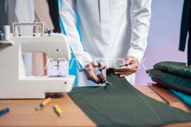 استعمال أدوات ومعدات الخياطة، حرف ومهن يدوية، إتقان صنع الملابس السعودية، بورتريه مقرب لخياط عربي خليجي سعودي يرتدي الثوب التقليدي يمسك المقص ويقص القماش، اختيار أجود أنواع الأقمشة لصناعة الأثواب، خلفية ملونة