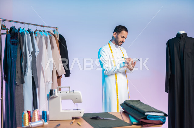 حرف ومهن يدوية، إتقان صنع الملابس السعودية، تسجيل وتدوين القياسات على الدفتر، بورتريه مقرب لخياط عربي خليجي سعودي يرتدي الثوب التقليدي يضع على رقبته الشريط المتري، استعمال أدوات ومعدات الخياطة، اختيار أجود أنواع الأقمشة لصناعة الأثواب، خلفية ملونة