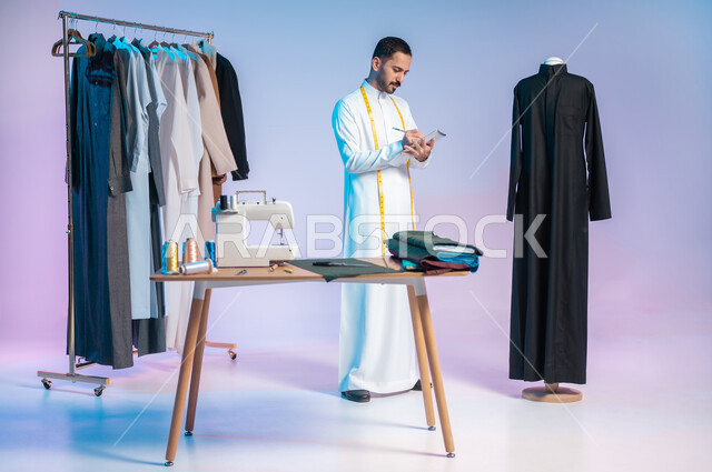 إتقان صنع الملابس السعودية، تسجيل وتدوين القياسات على الدفتر، بورتريه لخياط عربي خليجي سعودي يرتدي الثوب التقليدي يضع على رقبته الشريط المتري، استعمال أدوات ومعدات الخياطة، اختيار أجود أنواع الأقمشة لصناعة الأثواب، الجسم بالطول الكامل، خلفية ملونة