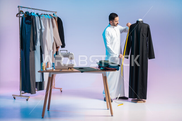 استعمال أدوات ومعدات الخياطة، استخدام الشريط المتري لأخذ القياسات، ترتيب وتنسيق الثياب، إتقان صنع الملابس السعودية، اختيار أجود أنواع الأقمشة لصناعة الأثواب، بورتريه لخياط عربي خليجي سعودي يرتدي الثوب التقليدي، مهن وحرف يدوية، الجسم بالطول الكامل، خلفية ملونة