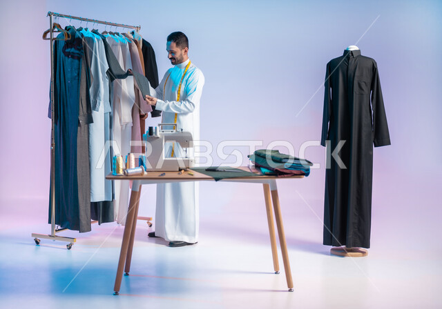 Practicing the profession of sewing, arranging and coordinating clothes, using sewing tools and equipment, mastering the manufacture of Saudi clothes, choosing the finest types of fabrics for making dresses, a portrait of a Saudi Arabian Gulf tailor wearing a traditional dress with a metric tape around his neck, full length body, colored background.