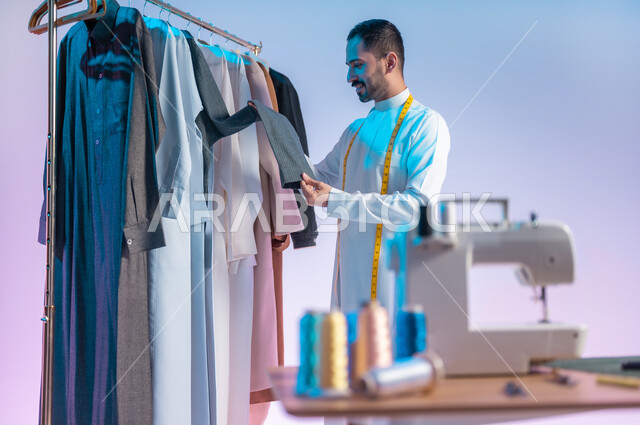 Arranging and coordinating clothes, using sewing tools and equipment, mastering the manufacture of Saudi clothes, choosing the finest types of fabrics for making dresses, a portrait of a Saudi Arabian Gulf tailor wearing a traditional dress with a metric tape around his neck to take measurements, professions and handicrafts, a colored background.