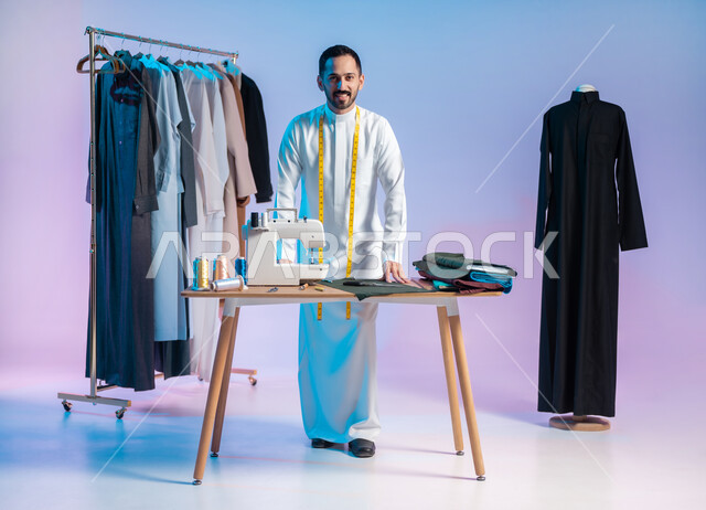 إتقان صنع الملابس السعودية، اختيار أجود أنواع الأقمشة لصناعة الأثواب، النظر للكاميرا بإيماءات السعادة، استعمال أدوات ومعدات الخياطة، بورتريه لخياط عربي خليجي سعودي يرتدي الثوب التقليدي يضع على رقبته الشريط المتري لأخذ القياسات، الجسم بالطول الكامل، خلفية ملونة