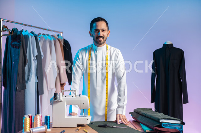اختيار أجود أنواع الأقمشة لصناعة الأثواب، مهن وحرف يدوية، النظر للكاميرا بإيماءات السعادة، إتقان صنع الملابس السعودية، استعمال أدوات ومعدات الخياطة، بورتريه مقرب لخياط عربي خليجي سعودي يرتدي الثوب التقليدي يضع على رقبته الشريط المتري لأخذ القياسات، خلفية ملونة