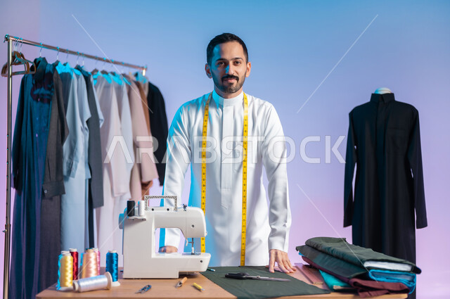 مهن وحرف يدوية، النظر للكاميرا بإيماءات السعادة، إتقان صنع الملابس السعودية، اختيار أجود أنواع الأقمشة لصناعة الأثواب، استعمال أدوات ومعدات الخياطة، بورتريه مقرب لخياط عربي خليجي سعودي يرتدي الثوب التقليدي يضع على رقبته الشريط المتري لأخذ القياسات، خلفية ملونة