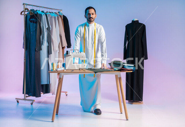 Looking at the camera with happy gestures, mastering the making of Saudi clothes, choosing the finest types of fabrics to make dresses, using sewing tools and equipment, a portrait of a Saudi Arabian Gulf tailor wearing a traditional dress with a metric tape around his neck to take measurements, the body in full length, a colored background.