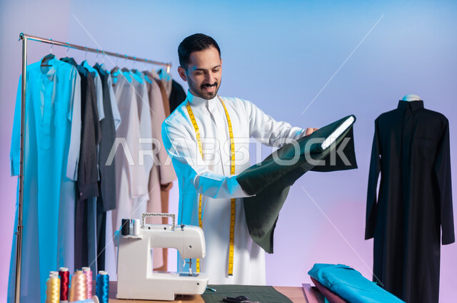 استعمال أدوات ومعدات الخياطة، اختيار أجود أنواع الأقمشة لصناعة الأثواب، إتقان صنع الملابس السعودية، بورتريه مقرب لخياط عربي خليجي سعودي يرتدي الثوب التقليدي يضع على رقبته الشريط المتري لأخذ القياسات، خلفية ملونة