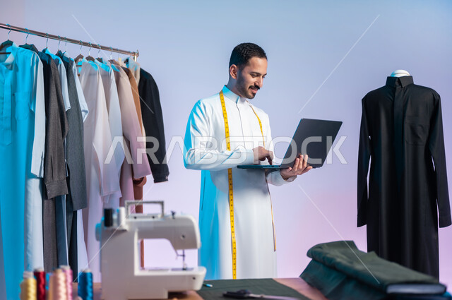 استخدام التكنولوجيا الحديثة والمتطورة، القيام بابتكار تصاميم وموديلات رائعة، استعمال أدوات ومعدات الخياطة، إتقان صنع الملابس السعودية، بورتريه مقرب لخياط عربي خليجي سعودي يرتدي الثوب التقليدي يستخدم الحاسب المحمول، خلفية ملونة