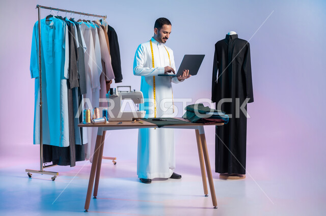 Creating wonderful designs and models, using modern and advanced technology, using sewing tools and equipment, mastering the making of Saudi clothes, a portrait of a Saudi Arabian Gulf tailor wearing a traditional dress, using a laptop, full-length body, colored background.