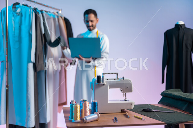 مهن وحرف يدوية، استخدام التقنيات المتطورة والحديثة، استعمال أدوات ومعدات الخياطة، إتقان صنع الملابس السعودية، بورتريه مقرب لخياط عربي خليجي سعودي يرتدي الثوب التقليدي يستخدم الحاسب المحمول، خلفية ملونة