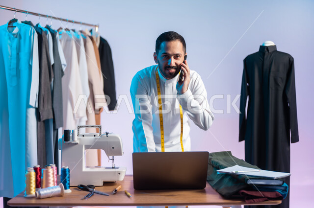 النظر للكاميرا بتعابير الفرح، تبادل الأخبار والأحاديث، إجراء مكالمة هاتفية، استخدام التقنيات المتطورة، استعمال أدوات ومعدات الخياطة، إتقان صنع الملابس السعودية، بورتريه مقرب لخياط عربي خليجي سعودي يرتدي الثوب التقليدي يستخدم الهاتف والحاسب المحمول، خلفية ملونة