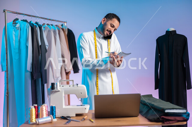 Recording and taking notes and measurements, making a phone call, using modern and advanced technologies, using sewing tools and equipment, mastering making Saudi clothes, a close-up portrait of a Saudi Arabian Gulf tailor wearing a traditional dress, using a mobile phone, a colorful background.