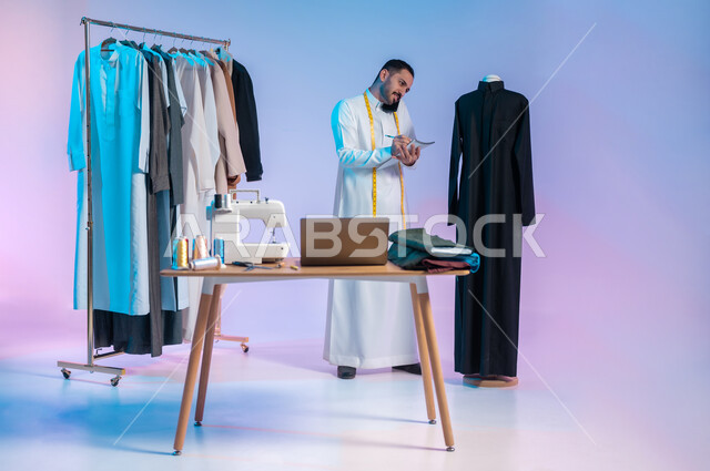تدوين الملاحظات و القياسات، إجراء مكالمة هاتفية، استخدام التقنيات الحديثة والمتطورة، استعمال أدوات ومعدات الخياطة، إتقان صنع الملابس السعودية، بورتريه لخياط عربي خليجي سعودي يرتدي الثوب التقليدي يستخدم  الهاتف المحمول، الجسم بالطول الكامل، خلفية ملونة