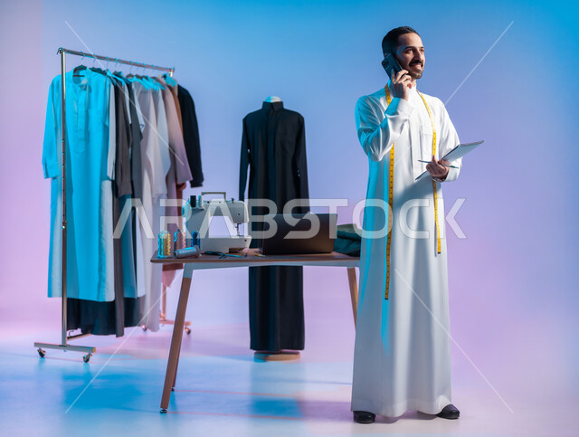 تعابير السعادة والسرور، إجراء مكالمة هاتفية، استخدام التقنيات الحديثة والمتطورة، استعمال أدوات ومعدات الخياطة، إتقان صنع الملابس السعودية، بورتريه لخياط عربي خليجي سعودي يرتدي الثوب التقليدي يستخدم  الهاتف المحمول، الجسم بالطول الكامل، خلفية ملونة