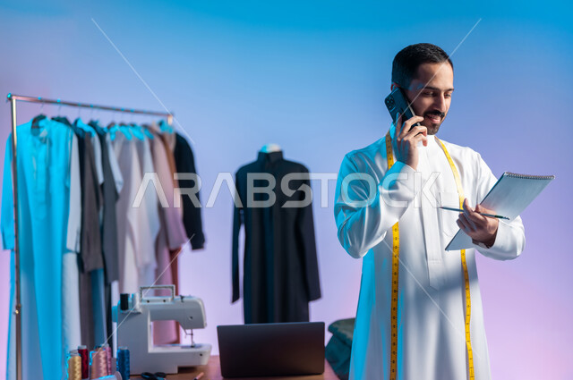 النظر الى الدفتر بإيماءات السعادة، إجراء مكالمة هاتفية، استخدام التقنيات الحديثة والمتطورة، استعمال أدوات ومعدات الخياطة، إتقان صنع الملابس السعودية، بورتريه مقرب لخياط عربي خليجي سعودي يرتدي الثوب التقليدي يستخدم  الهاتف المحمول، خلفية ملونة