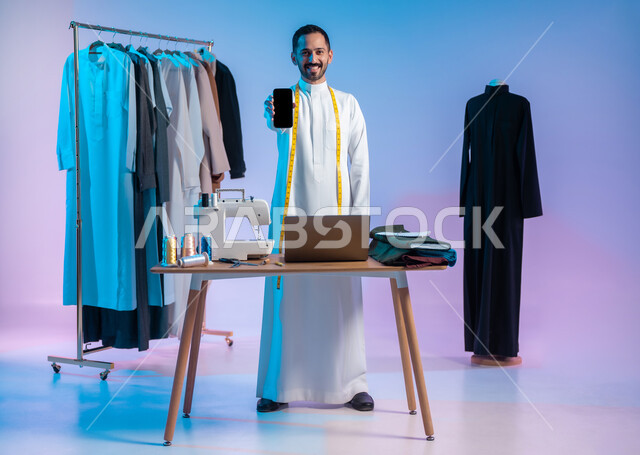 Using advanced technologies, looking at the camera with gestures of happiness and pleasure, using sewing tools and equipment, mastering the making of Saudi clothes, a portrait of a Saudi Arabian Gulf tailor wearing a traditional dress, using a mobile phone, displaying a blank black screen, the body full length, colored background.