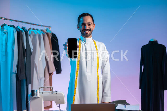 النظر الى الكاميرا بإيماءات السعادة والسرور، استخدام التقنيات الحديثة والمتطورة، استعمال أدوات ومعدات الخياطة، إتقان صنع الملابس السعودية، بورتريه مقرب لخياط عربي خليجي سعودي يرتدي الثوب التقليدي يستخدم  الهاتف المحمول، عرض شاشة سوداء فارغة، خلفية ملونة