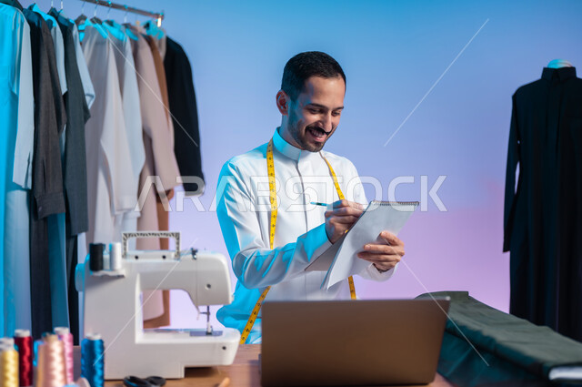 العمل في مهنة الخياطة، تصميم ورسم الأزياء على الدفتر، استخدام التكنولوجيا الحديثة والمتطورة، استعمال أدوات ومعدات الخياطة، إتقان صنع الملابس السعودية، بورتريه مقرب لخياط عربي خليجي سعودي يرتدي الثوب التقليدي يستخدم الحاسب المحمول، خلفية ملونة