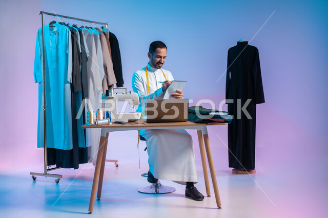تصميم ورسم الأزياء على الدفتر، استخدام التكنولوجيا الحديثة والمتطورة، استعمال أدوات ومعدات الخياطة، إتقان صنع الملابس السعودية، بورتريه لخياط عربي خليجي سعودي يرتدي الثوب التقليدي يستخدم الحاسب المحمول، الجسم بالطول الكامل، خلفية ملونة