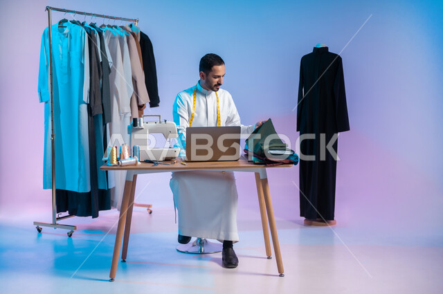 اختيار أجود أنواع الأقمشة لصناعة الأثواب، استخدام التكنولوجيا الحديثة والمتطورة، استعمال أدوات ومعدات الخياطة، إتقان صنع الملابس السعودية، بورتريه لخياط عربي خليجي سعودي يرتدي الثوب التقليدي يستخدم الحاسب المحمول، الجسم بالطول الكامل، خلفية ملونة