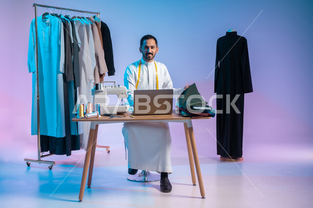 استخدام التكنولوجيا الحديثة والمتطورة، النظر الى الكاميرا بإيماءات السعادة، استعمال أدوات ومعدات الخياطة، إتقان صنع الملابس السعودية، بورتريه لخياط عربي خليجي سعودي يرتدي الثوب التقليدي يستخدم الحاسب المحمول، الجسم بالطول الكامل، خلفية ملونة