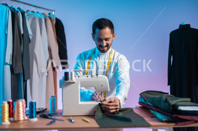 إتقان صنع الملابس السعودية، استخدام ماكينة الخياطة لتفصيل الثياب، بورتريه مقرب لخياط عربي خليجي سعودي يرتدي الثوب التقليدي يقوم بصناعة الأثواب، استعمال أدوات ومعدات الخياطة، خلفية ملونة