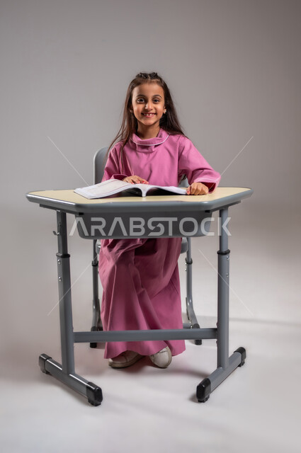 أهمية العلم في تنمية القدرات العقلية، العودة إلى المدارس والطريق الى النجاح،  بورتريه لطالبة عربية سعودية خليجية جالسة على المقعد المدرسي تستعد لتتلقى المعلومات من مدرستها، ايماءات الفرح و الحماس بقدوم الفصل الدراسي، خلفية رمادية